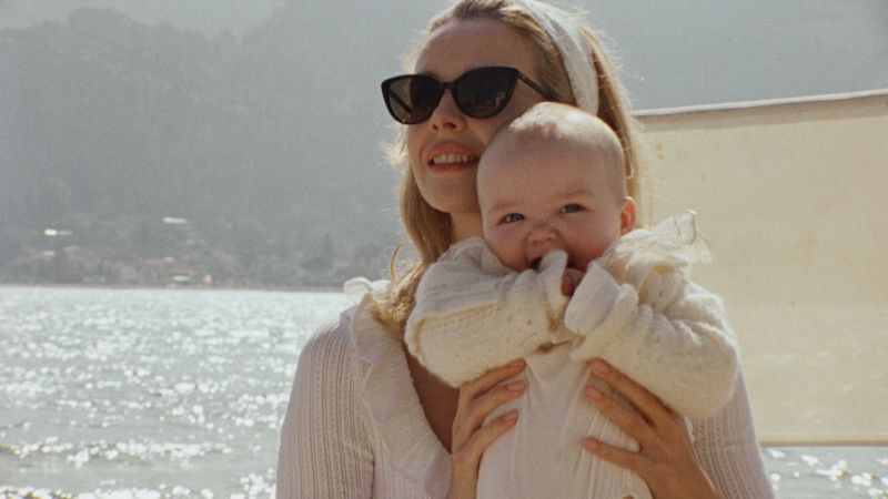 Greta Bellamacina wearing dark sunglasses and holding an infant dressed in an ivory knit outfit by the sea.