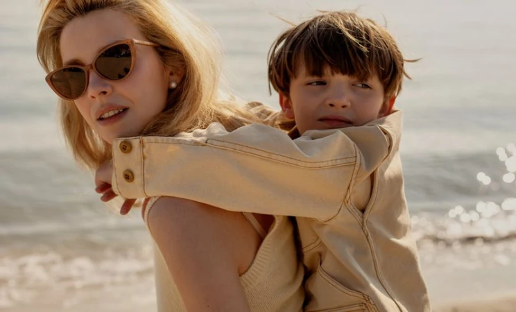 Greta Bellamacina wearing cat-eye sunglasses while carrying a young boy on her back at a sunny beach.