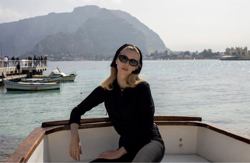 Model Greta Bellamacina wearing sunglasses and black attire seated in a boat on the water for the Doen x Garrett Leight campaign.