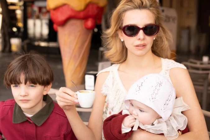 Greta Bellamacina wearing sunglasses and holding an espresso cup alongside two children in an outdoor café setting