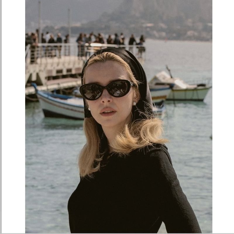 Model Greta Bellamacina wearing black cat-eye sunglasses and a black headscarf against a coastal harbor with boats.