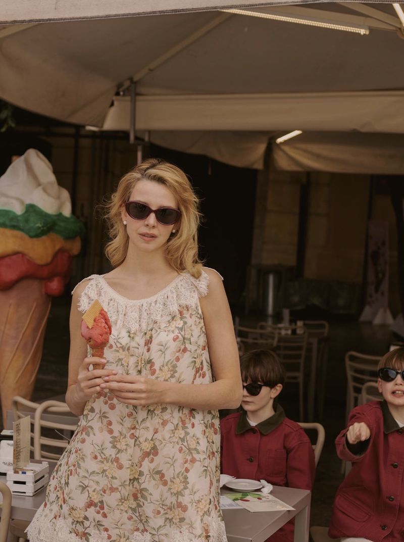 Model Greta Bellamacina wearing dark sunglasses and a floral sleeveless dress holding a gelato cone at an outdoor cafe table.