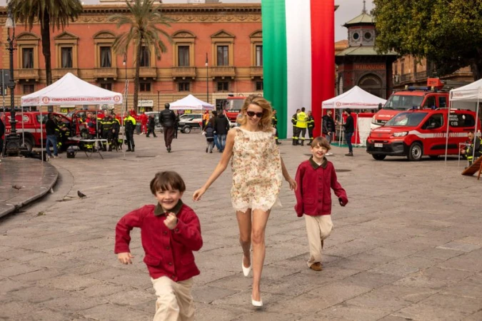 Greta Bellamacina in a floral mini dress runs across a public square with two boys in red jackets during a brand campaign shoot.