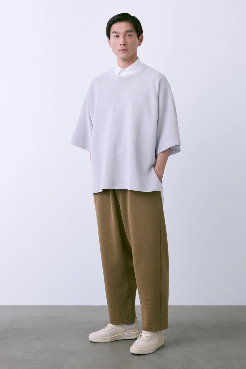 Studio shot of an adult male in casual fashion, wearing a light gray shirt, tan pants, and sneakers against a white background.