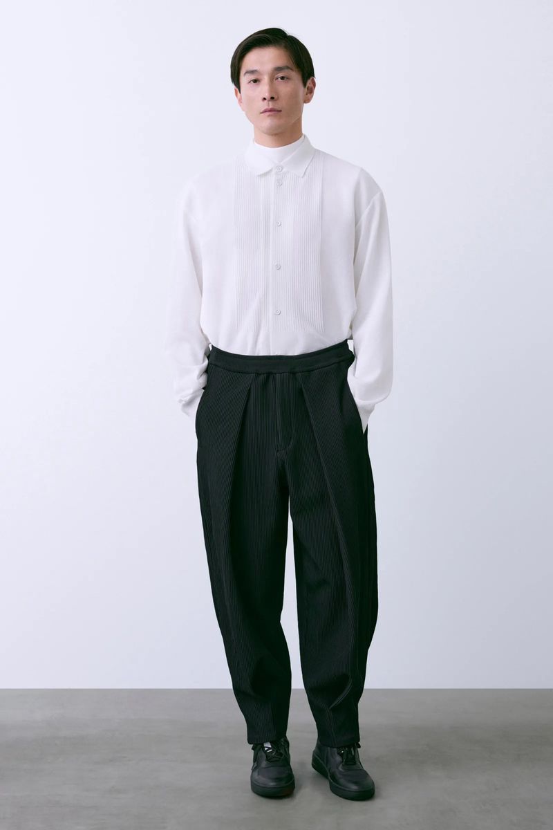 Full-length studio shot of an adult male wearing a white collared shirt and black pleated pants with black sneakers.