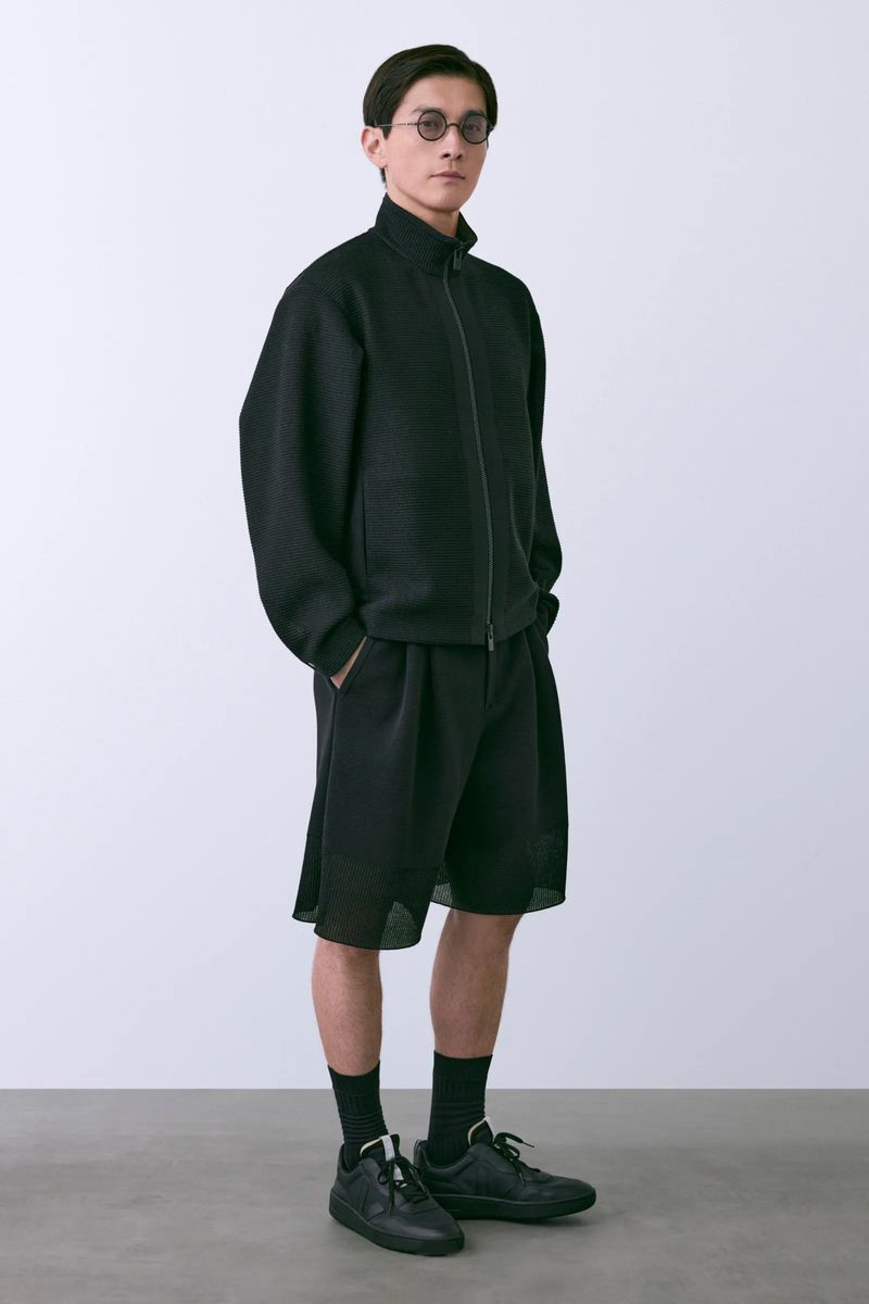 A slender Asian man in a black zip-up jacket, baggy shorts, and sneakers poses in a bright studio with hands in pockets.