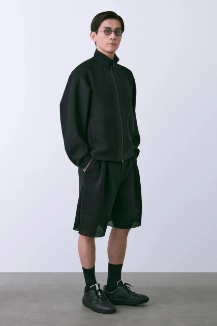 A slender Asian man in a black zip-up jacket, baggy shorts, and sneakers poses in a bright studio with hands in pockets.