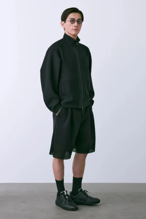A slender Asian man in a black zip-up jacket, baggy shorts, and sneakers poses in a bright studio with hands in pockets.