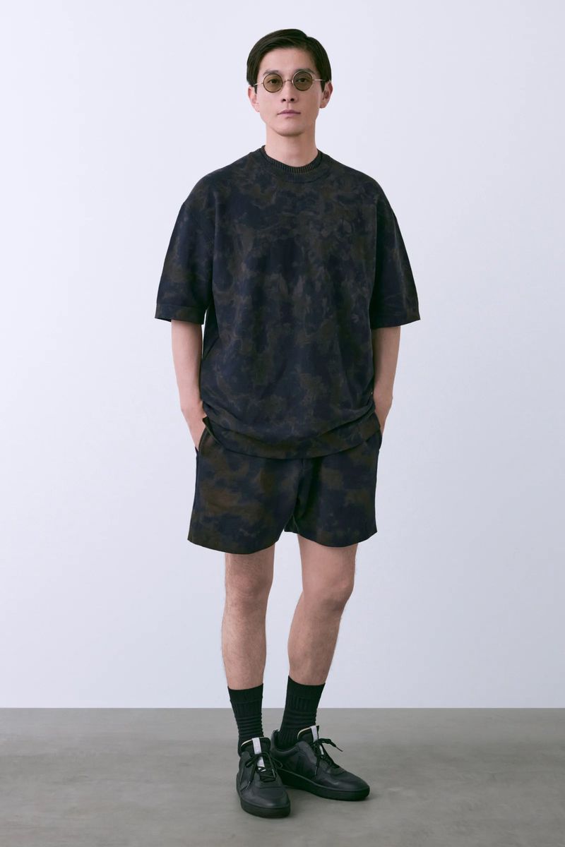 Model Daisuke Ueda in dark patterned t-shirt, matching shorts, sunglasses, and black sneakers against a white studio background.