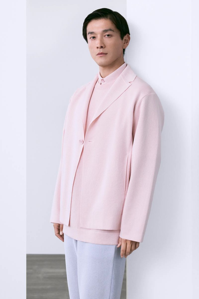 Portrait of a stylish adult male with dark hair wearing a pale pink blazer and trousers, posing against a bright white corner wall.
