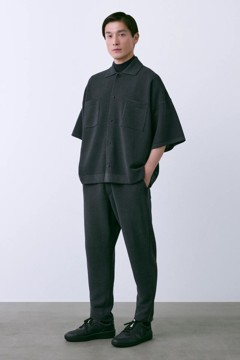Studio shot of an adult male model in a grey knit collared shirt, turtleneck, and pants, standing against a white background.
