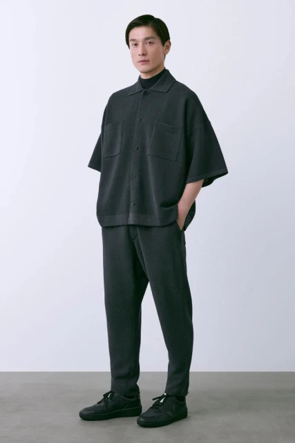 Studio shot of an adult male model in a grey knit collared shirt, turtleneck, and pants, standing against a white background.