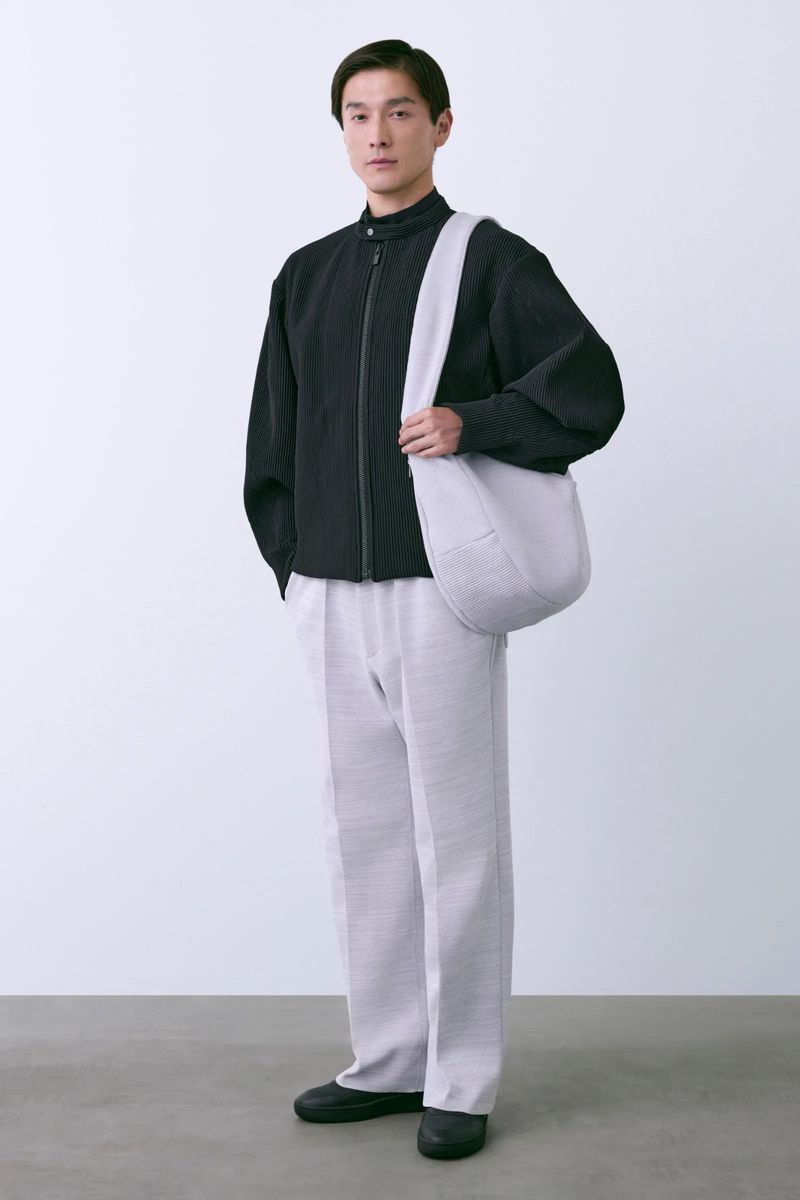 Studio shot of a male model wearing a black jacket, light gray pants, and carrying a light gray bag, posing against a white backdrop.
