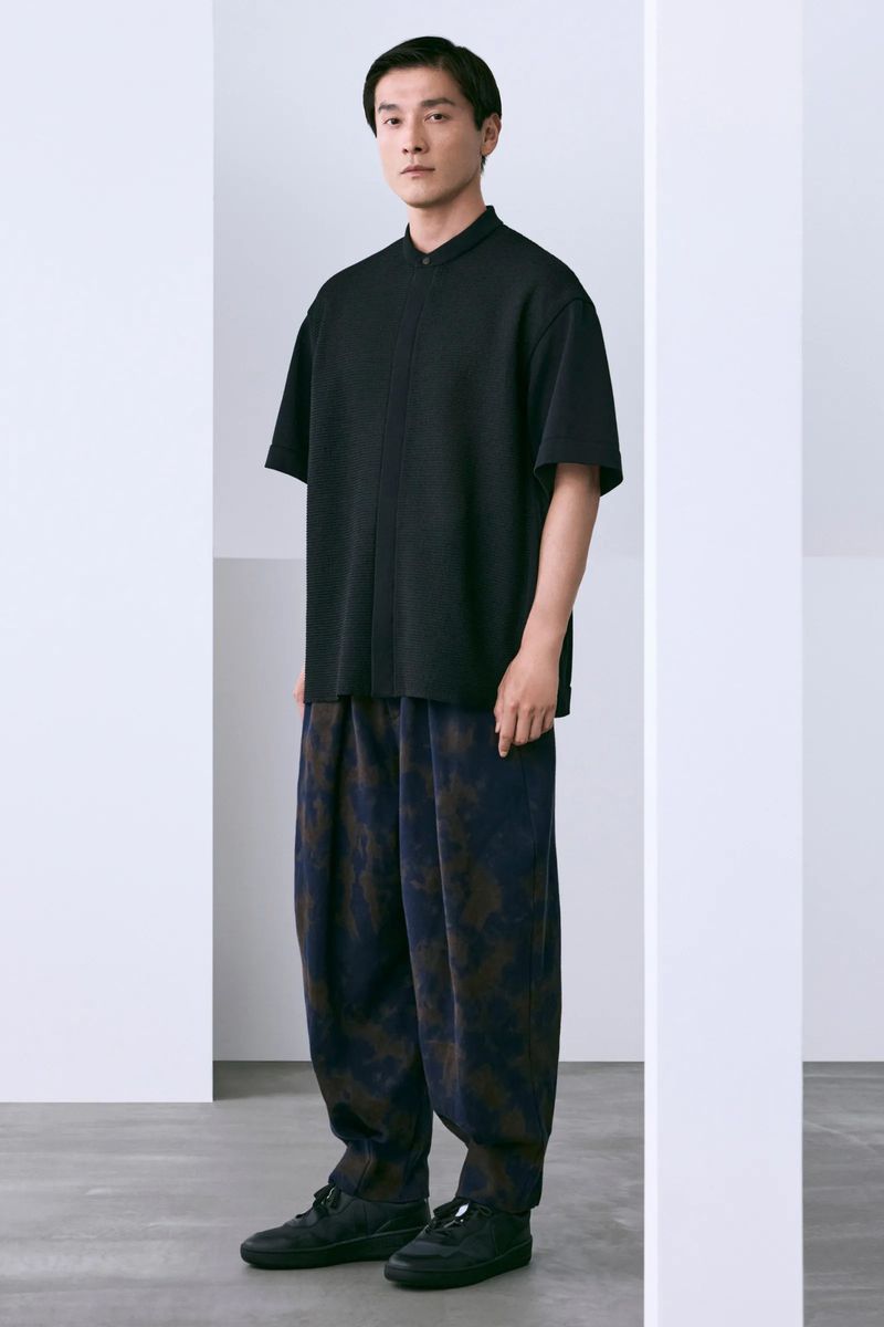 Model Daisuke Ueda stands framed between white architectural pillars wearing a black short-sleeved top and patterned wide trousers.