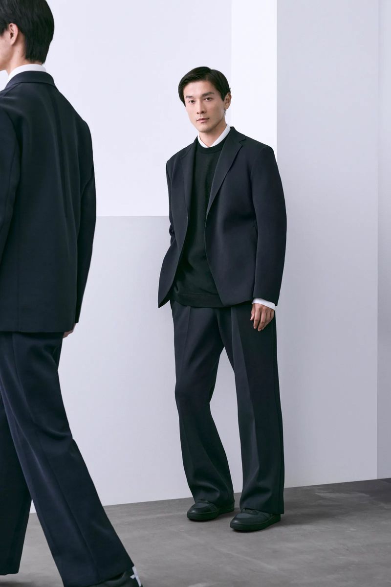 Man in a navy suit with loose pants stands in a bright studio. A second man, blurred in motion, is partially visible in the foreground.