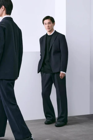 Man in a navy suit with loose pants stands in a bright studio. A second man, blurred in motion, is partially visible in the foreground.