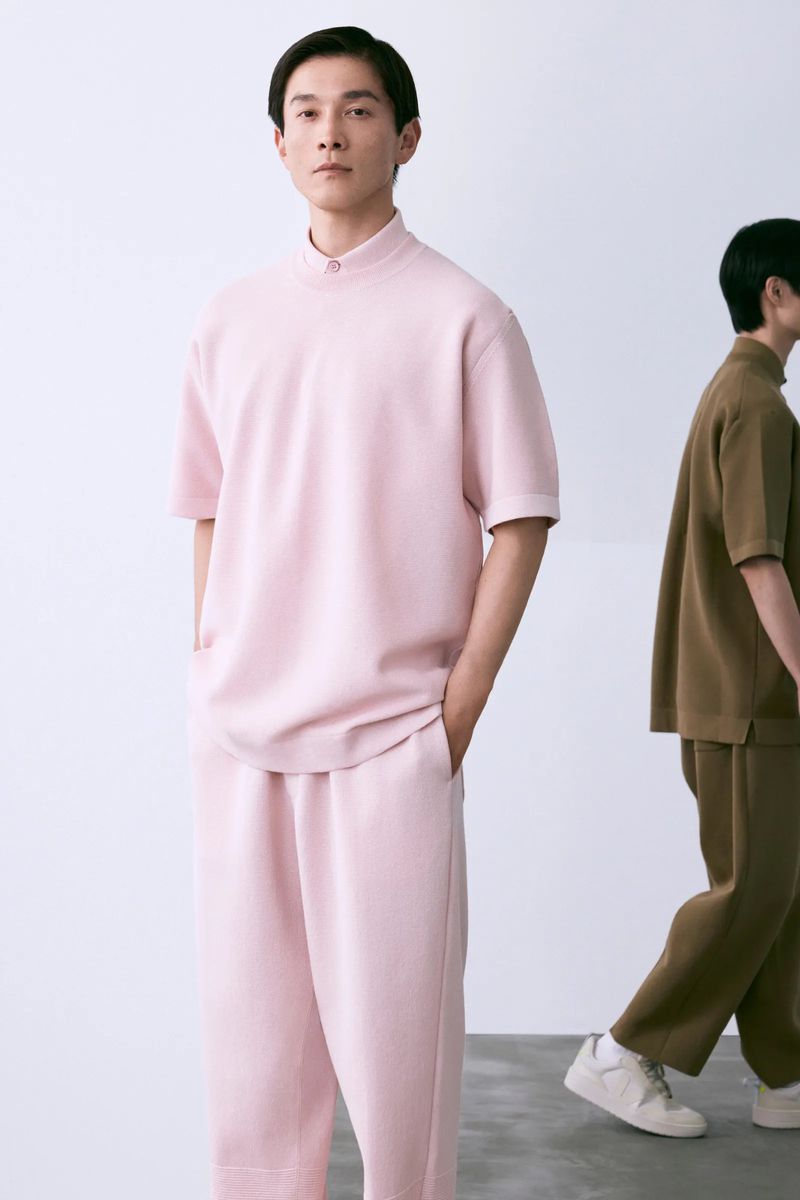 Two Asian men pose in matching outfits in a minimalist photo studio, one in pink, one in brown, with a clean white background.