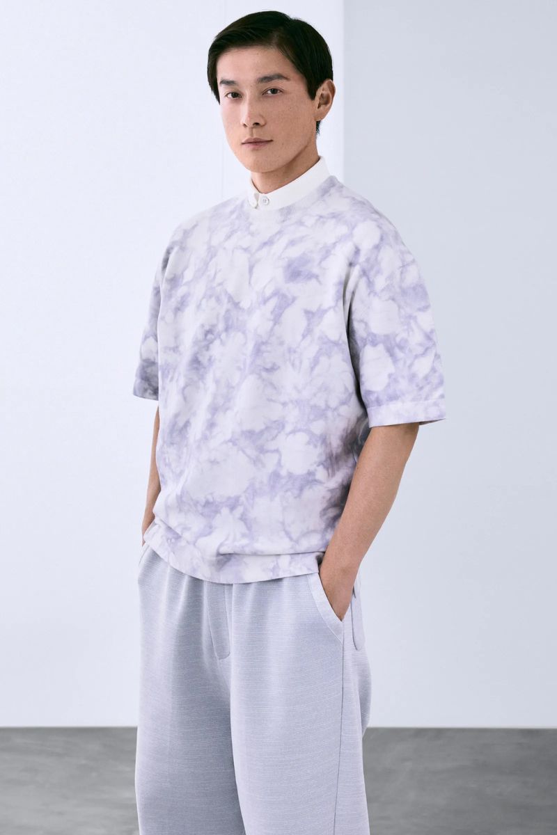 A young adult male stands indoors wearing a patterned grey t-shirt and matching pants, hands casually in his pockets. Soft, natural light.