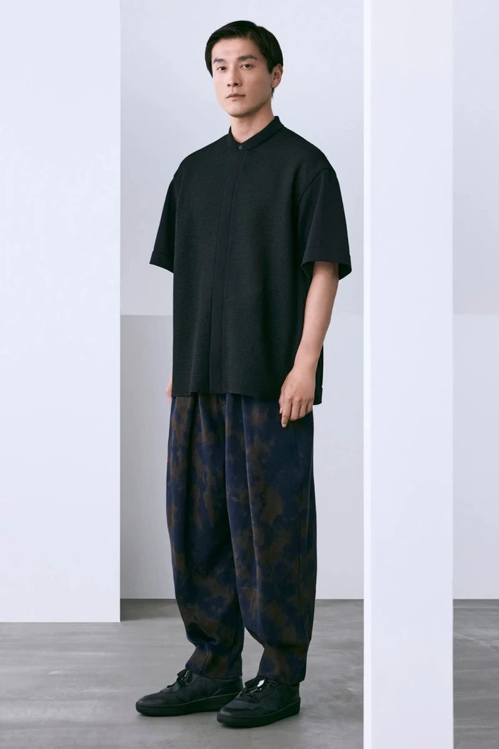 Model Daisuke Ueda stands framed between white architectural pillars wearing a black short-sleeved top and patterned wide trousers.
