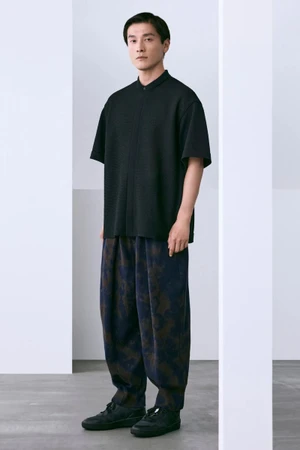 Model Daisuke Ueda stands framed between white architectural pillars wearing a black short-sleeved top and patterned wide trousers.