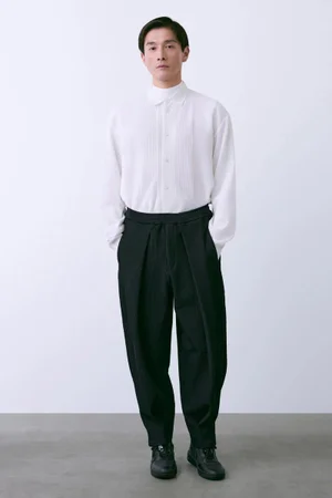 Model Daisuke Ueda in a white button-up shirt and black pleated trousers against a plain studio background for CFCL S/S 26.