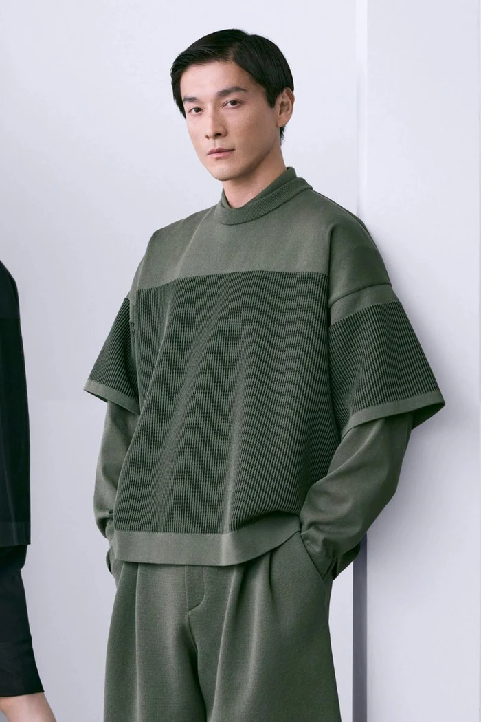 Asian male model wearing an olive green textured knit layered sweater and matching pants against a clean white studio wall.