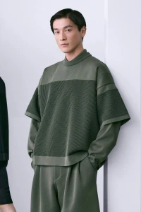 Asian male model wearing an olive green textured knit layered sweater and matching pants against a clean white studio wall.