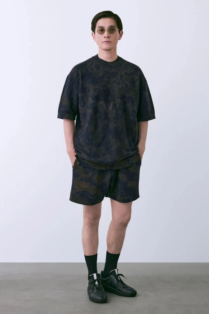 Model Daisuke Ueda in dark patterned t-shirt, matching shorts, sunglasses, and black sneakers against a white studio background.