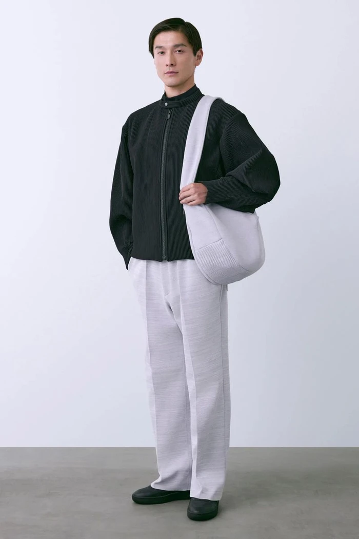 Asian male model wearing a dark zip jacket, light trousers, and carrying a shoulder bag against a white seamless background