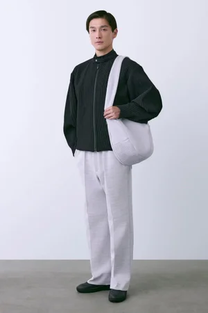 Asian male model wearing a dark zip jacket, light trousers, and carrying a shoulder bag against a white seamless background