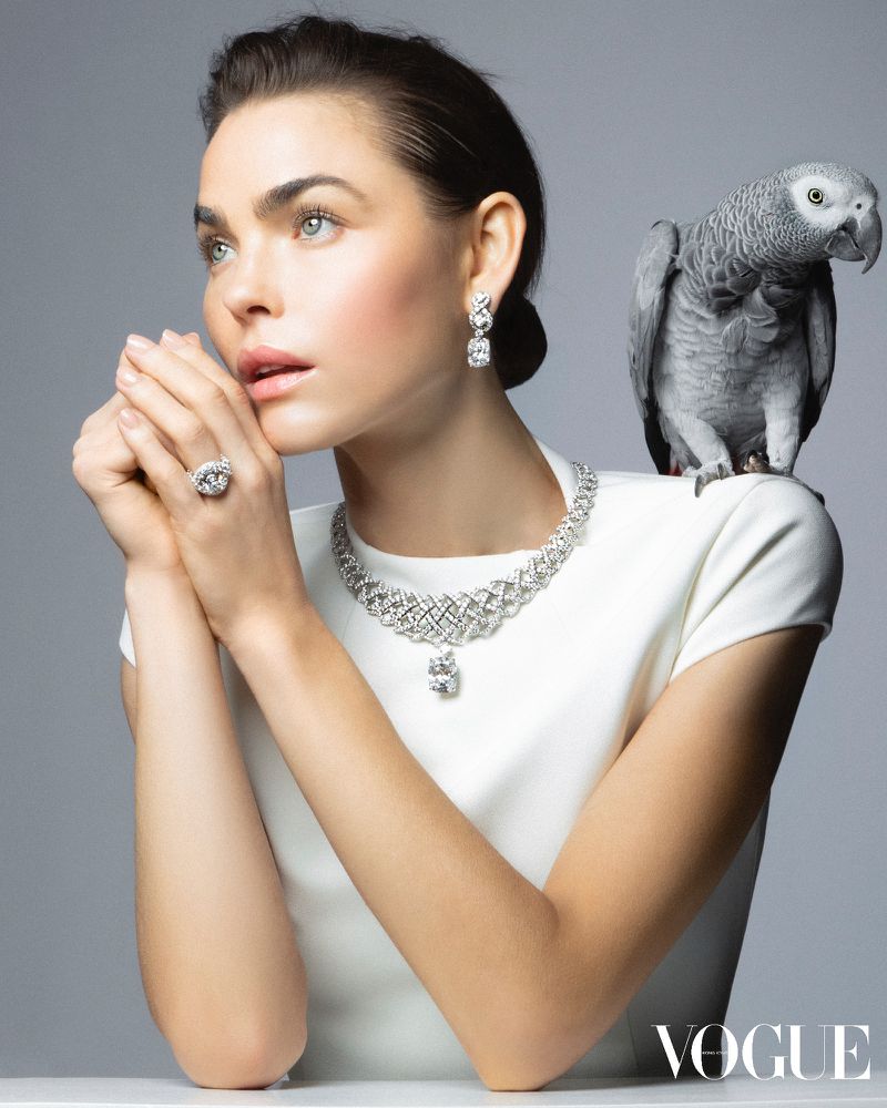 Model Bambi Northwood Blyth wearing diamond jewelry with an African grey parrot on her shoulder against a grey studio backdrop.