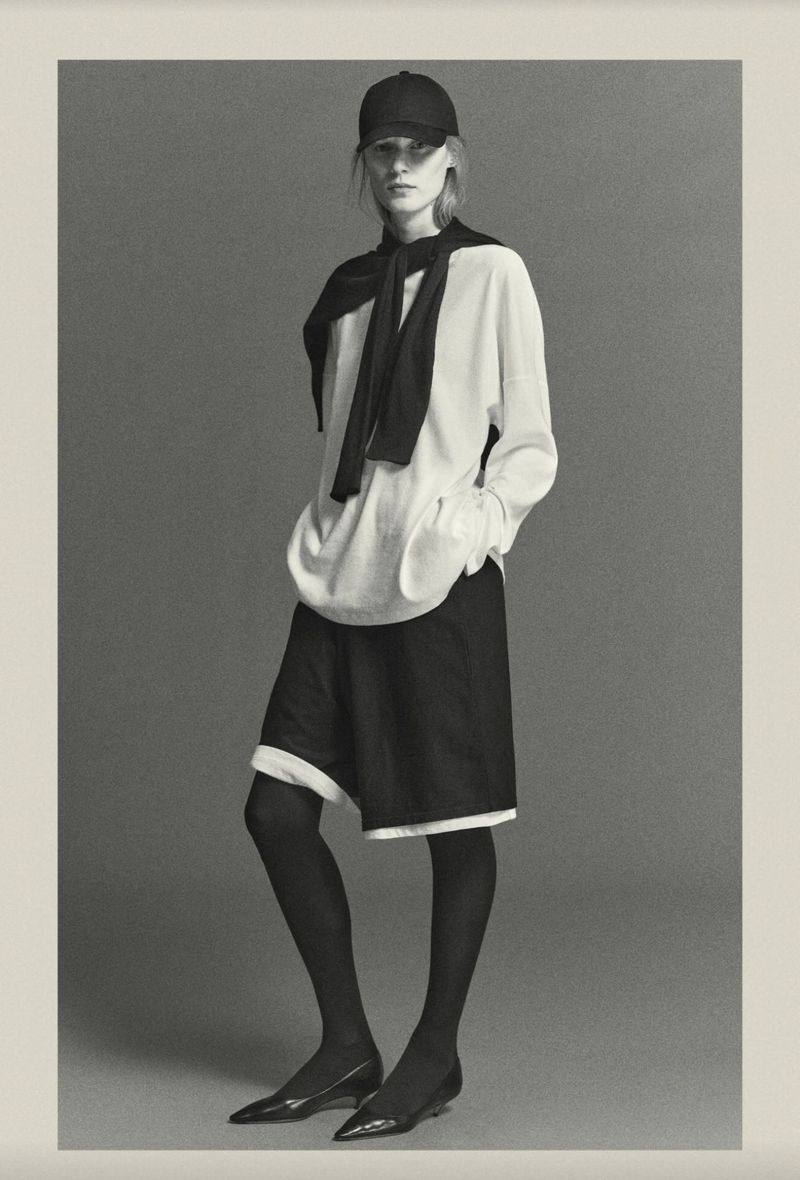 Fashion portrait of a woman wearing a white shirt, dark shorts, black tights, and a baseball cap in a photo studio setting.