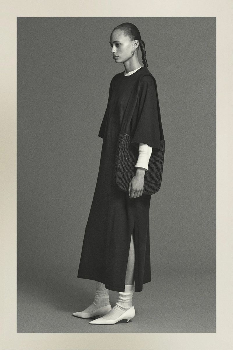 Monochrome studio shot of a woman in a black dress with side slit, accessorized with a woven bag, posing against a neutral background.