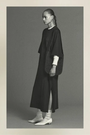 Monochrome studio shot of a woman in a black dress with side slit, accessorized with a woven bag, posing against a neutral background.