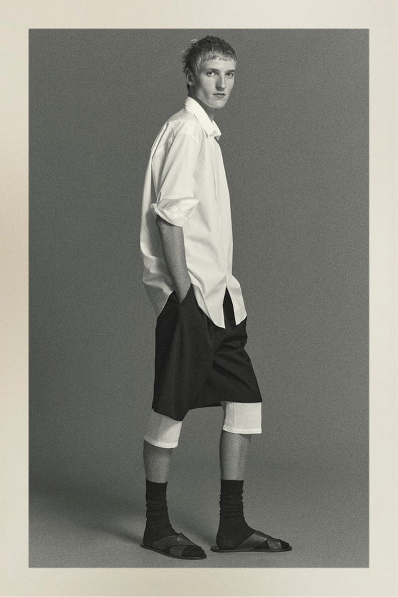 Black and white full-length studio shot of an adult male wearing a white button-down shirt, black shorts, socks, and sandals, hands in pockets.