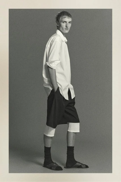 Black and white full-length studio shot of an adult male wearing a white button-down shirt, black shorts, socks, and sandals, hands in pockets.