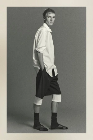 Black and white full-length studio shot of an adult male wearing a white button-down shirt, black shorts, socks, and sandals, hands in pockets.