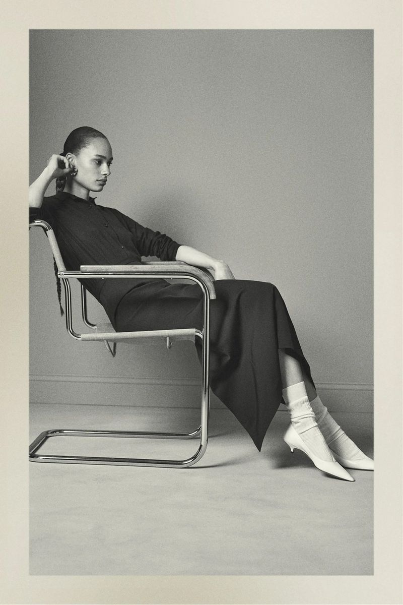 Elegant woman in a black dress sitting in a chrome chair in a photo studio. She has white socks and white shoes on.
