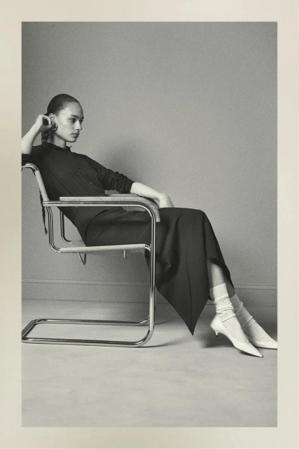 Elegant woman in a black dress sitting in a chrome chair in a photo studio. She has white socks and white shoes on.