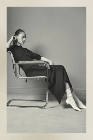 Elegant woman in a black dress sitting in a chrome chair in a photo studio. She has white socks and white shoes on.