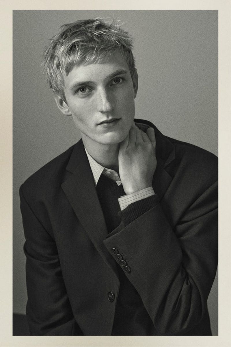 Black and white portrait of a young adult male model in a dark suit jacket, touching his neck in a photo studio setting.