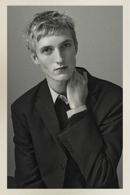 Black and white portrait of a young adult male model in a dark suit jacket, touching his neck in a photo studio setting.