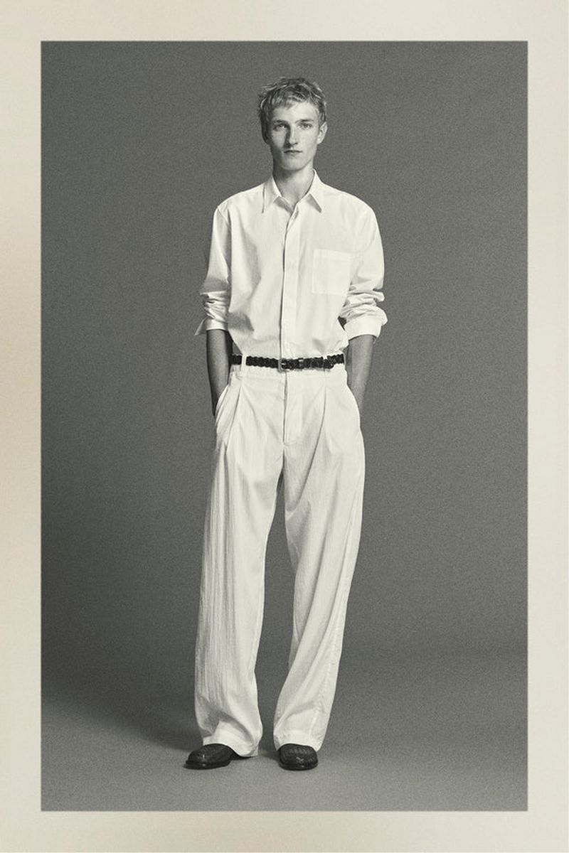 Full-length portrait of a young adult male in an unbuttoned white shirt and light-colored pants against a neutral grey backdrop.