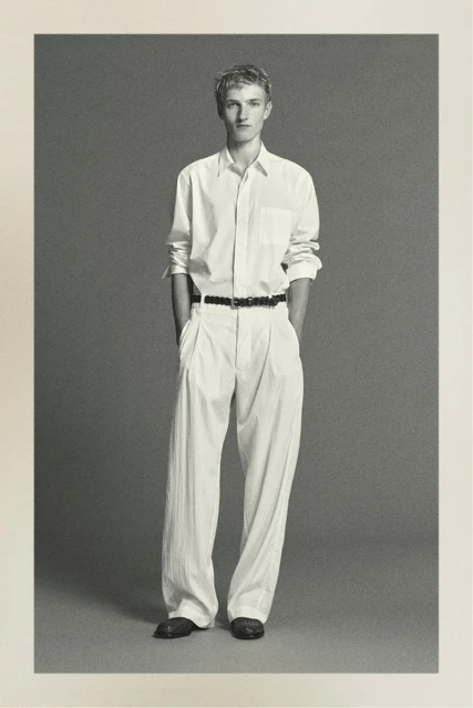 Full-length portrait of a young adult male in an unbuttoned white shirt and light-colored pants against a neutral grey backdrop.