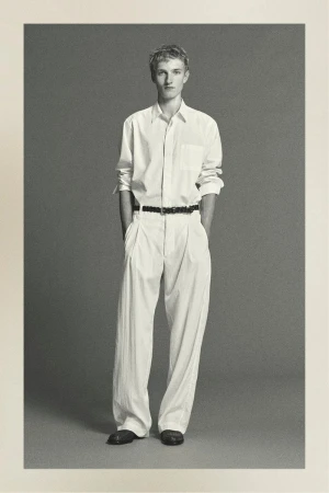 Full-length portrait of a young adult male in an unbuttoned white shirt and light-colored pants against a neutral grey backdrop.