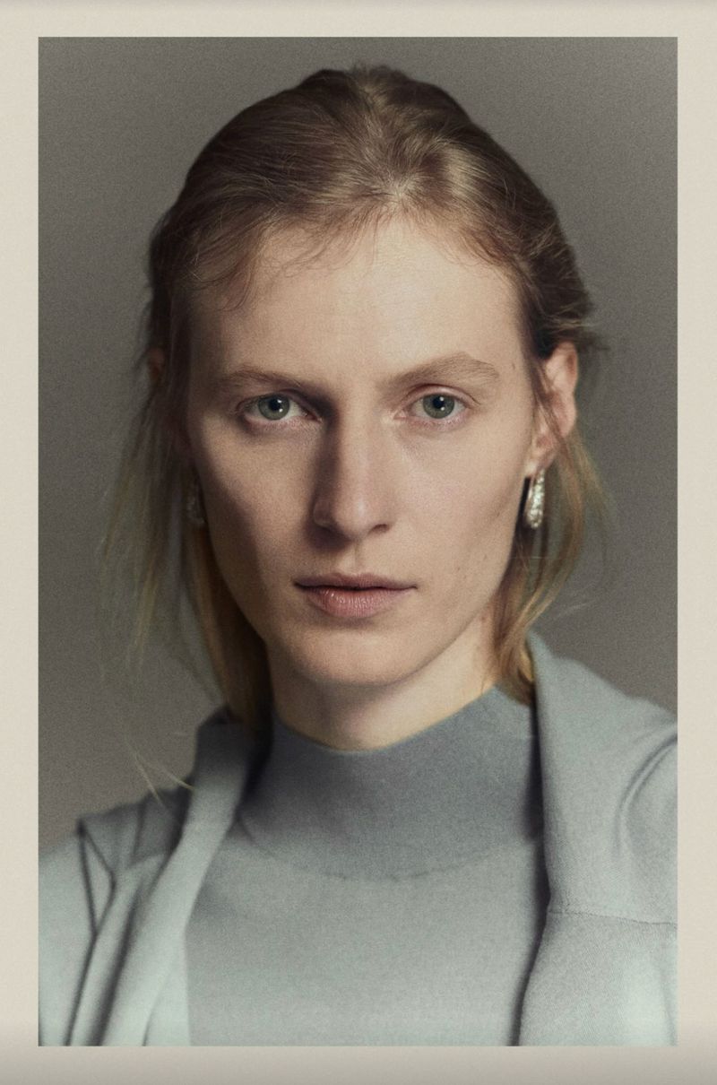 Portrait of a young adult female with blonde hair, wearing a blue turtleneck sweater and jacket in a neutral indoor setting.