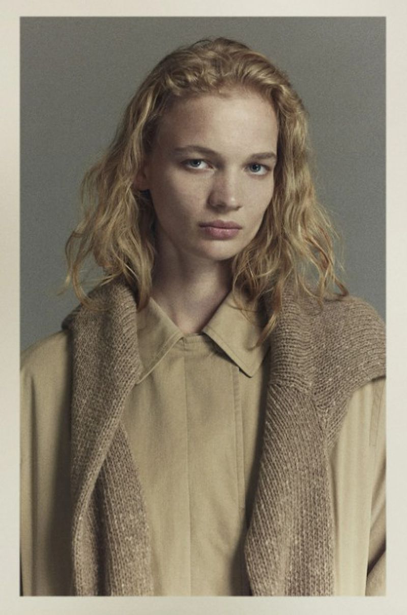 Blonde-haired woman with a neutral facial expression wearing a tan-colored collared shirt and sweater around her shoulders indoors.