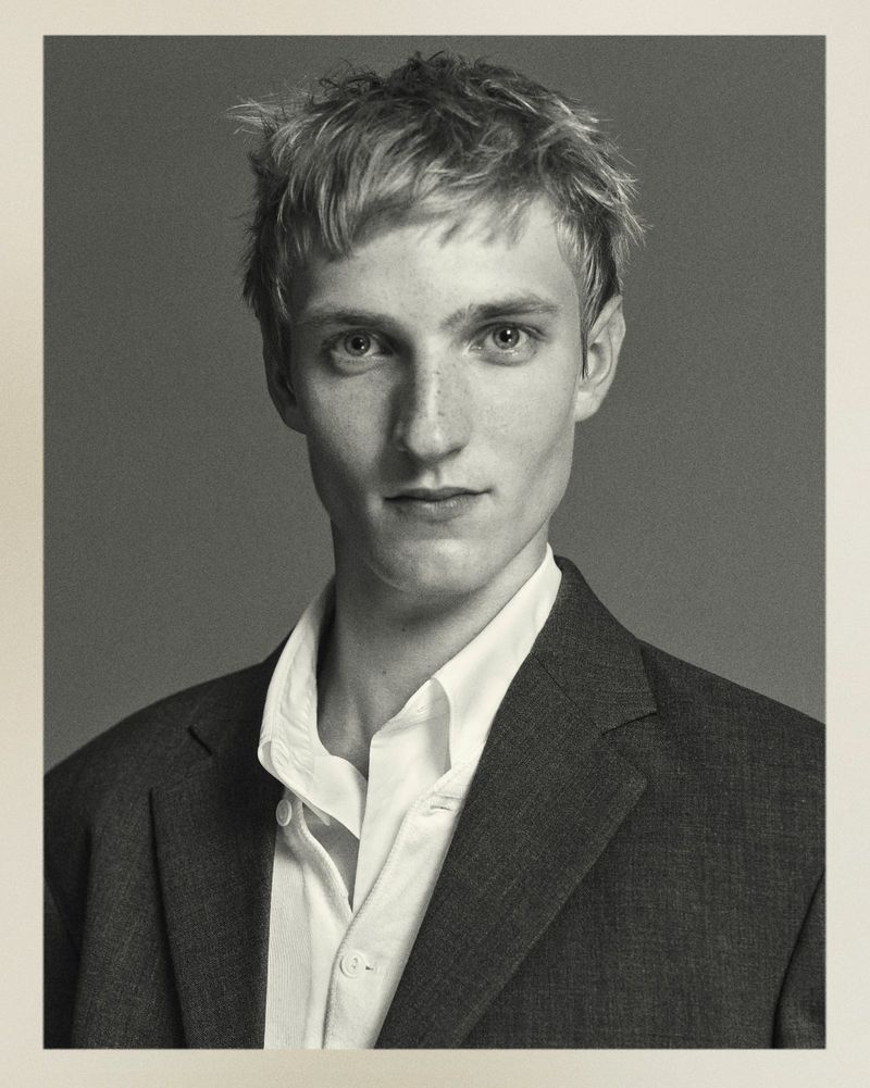 Black and white portrait of a young adult male with light hair in a dark suit jacket and white collared shirt against a grey background.