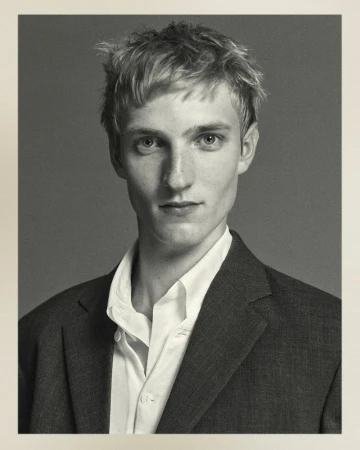 Black and white portrait of a young adult male with light hair in a dark suit jacket and white collared shirt against a grey background.