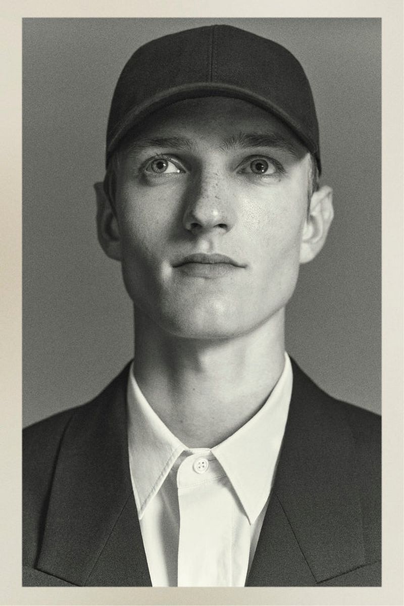 Portrait of a young fair-skinned adult male wearing a baseball cap, collared shirt, and blazer, set against a muted grey backdrop.